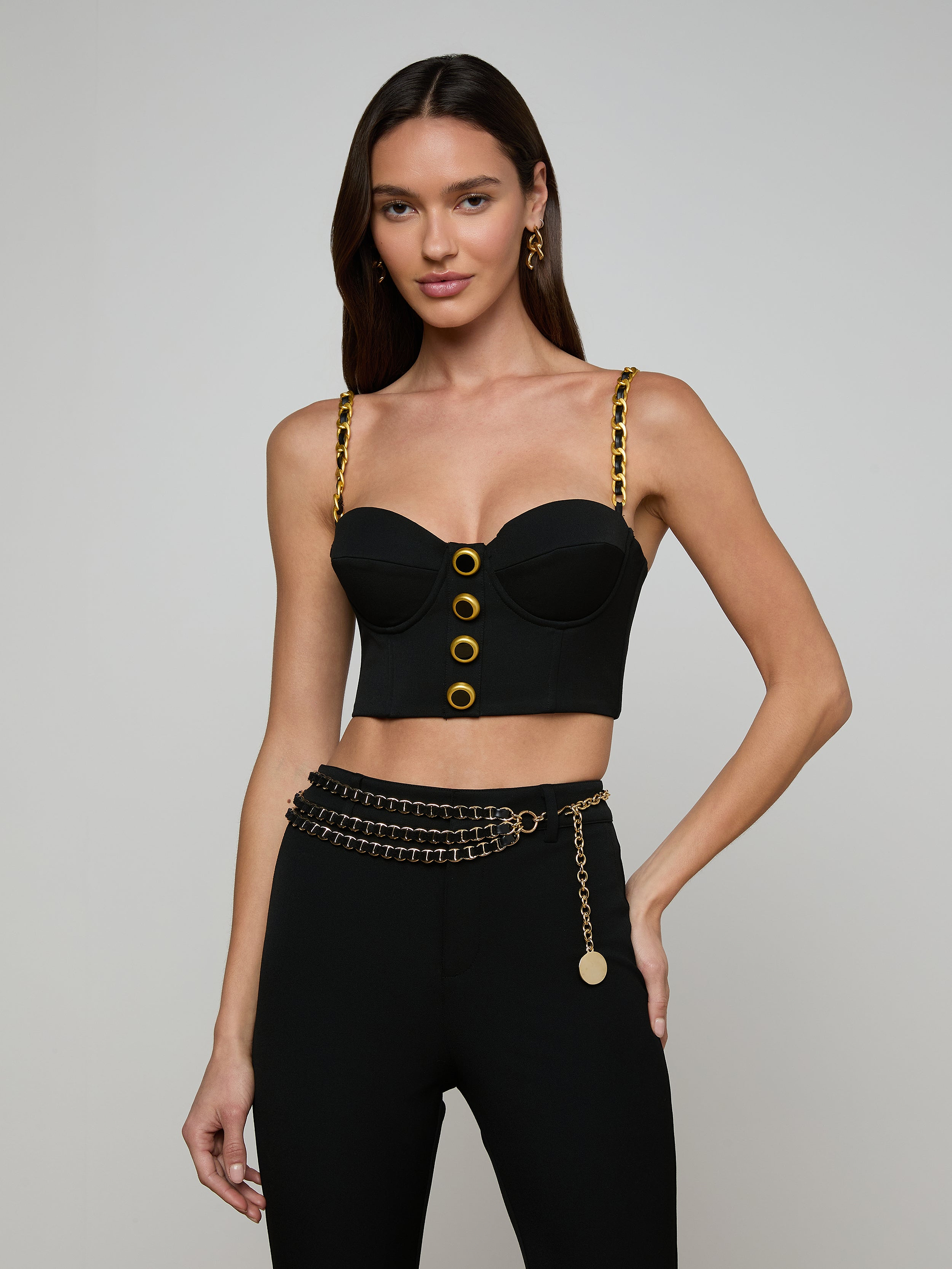 Wearing the Sophia Chain Bralette—a black crop top with gold chain straps and button accents that creates a structured silhouette—she pairs it with high-waisted black pants in stretch suiting fabric, finished with a gold chain belt, standing against a light background.