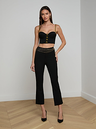 A woman wearing a black Sophia Chain Bralette with gold straps and buttons, black pants crafted from stretch suiting fabric with a gold chain belt, and black heels stands against a plain white background.