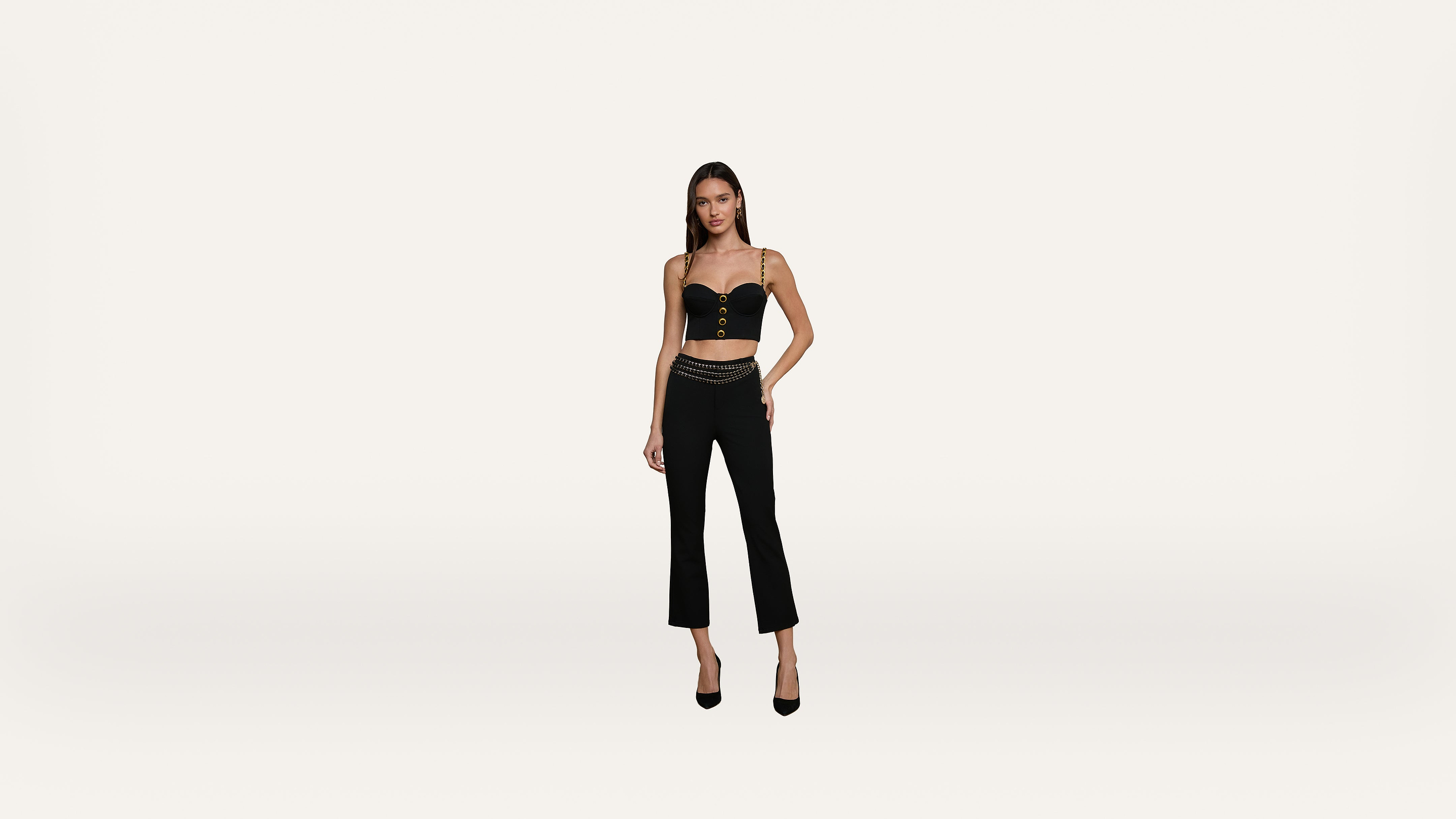 A woman wearing a black crop top, black high-waisted pants, and black heels stands against a plain white background, with one hand on her hip.