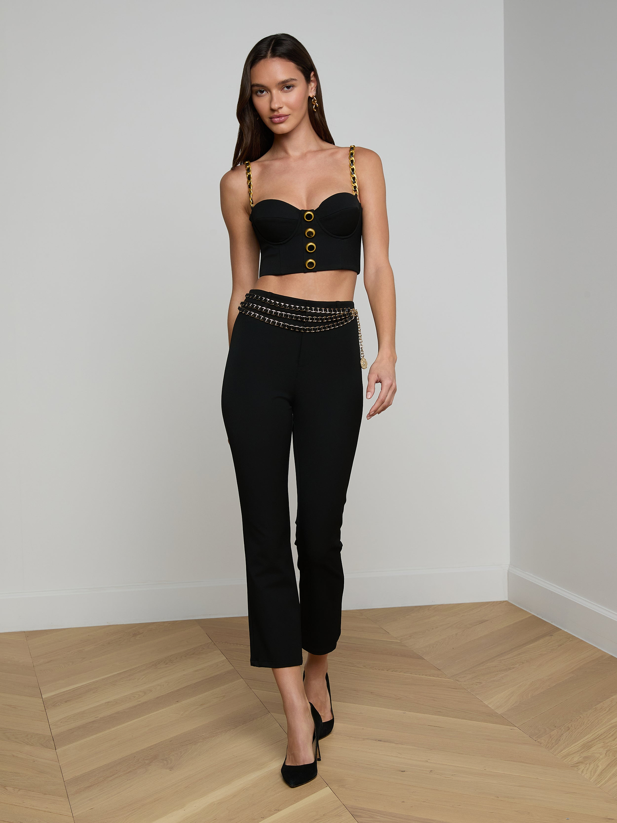 A woman wearing a black Sophia Chain Bralette with gold chain details and black pants, walking forward against a plain light background, paired with black high-heeled shoes. The outfit features a structured silhouette crafted from stretch suiting fabric.