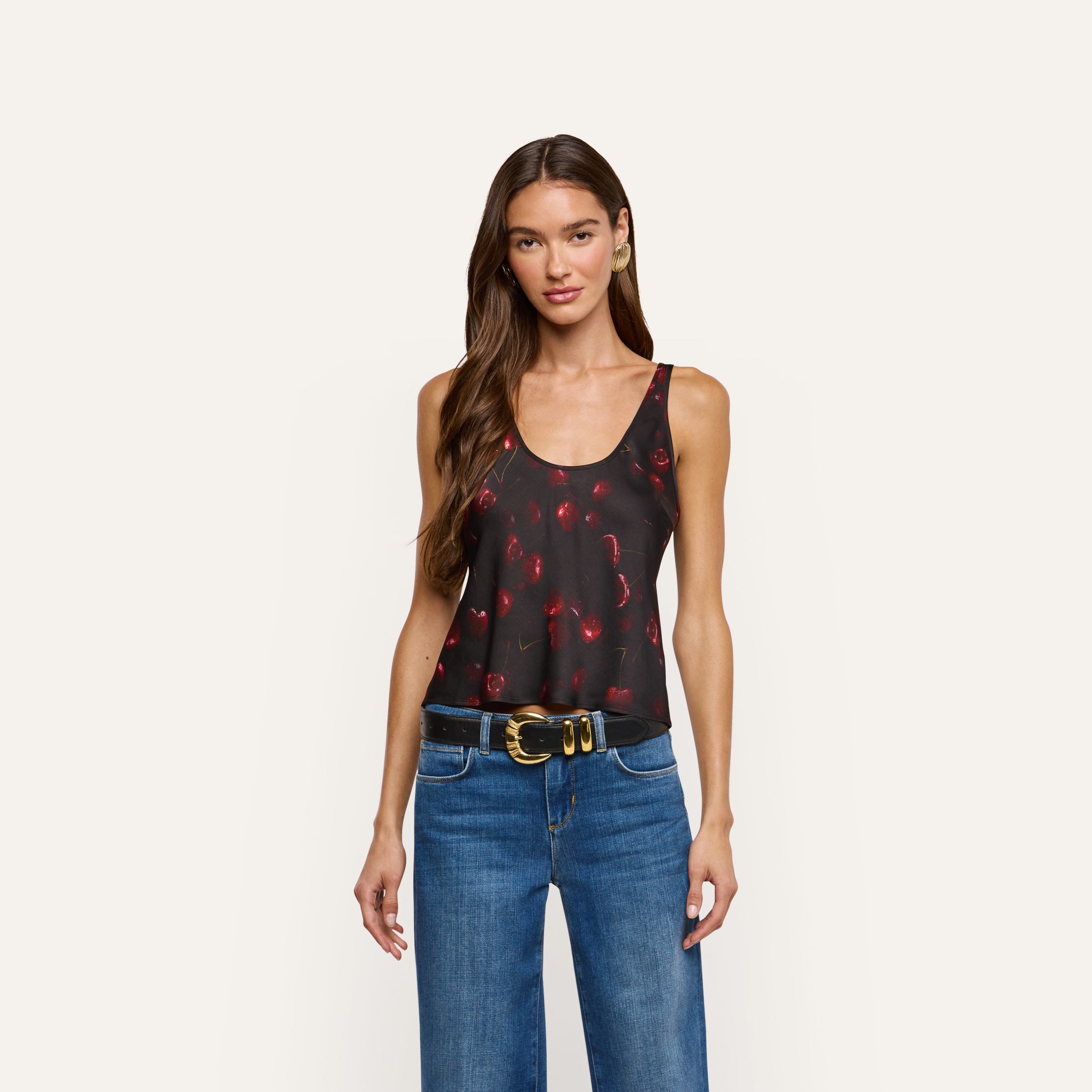 A person wearing an Eddie Satin Tank in black with a red cherry print, blue jeans, and a black belt.