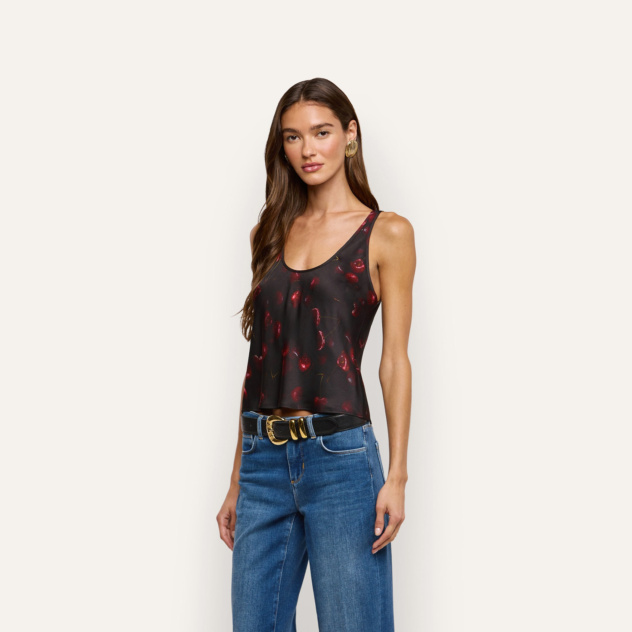 The Eddie Satin Tank, a black bias-cut satin top with a cherry print, worn tucked into blue jeans with a black belt featuring a gold buckle.