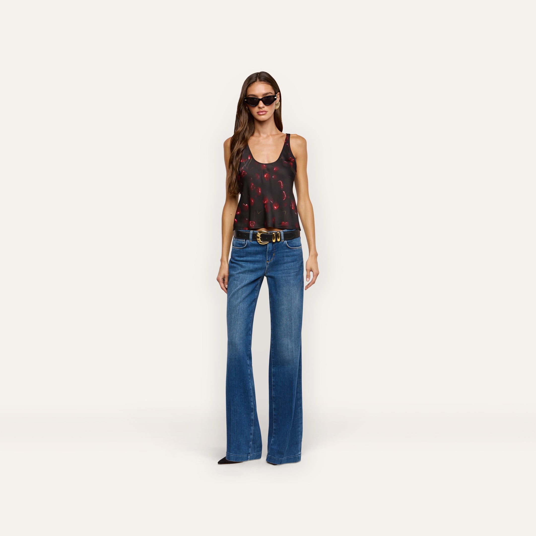 A model wears an Eddie Satin Tank with a red cherry print, tucked into blue flared jeans with a black belt.