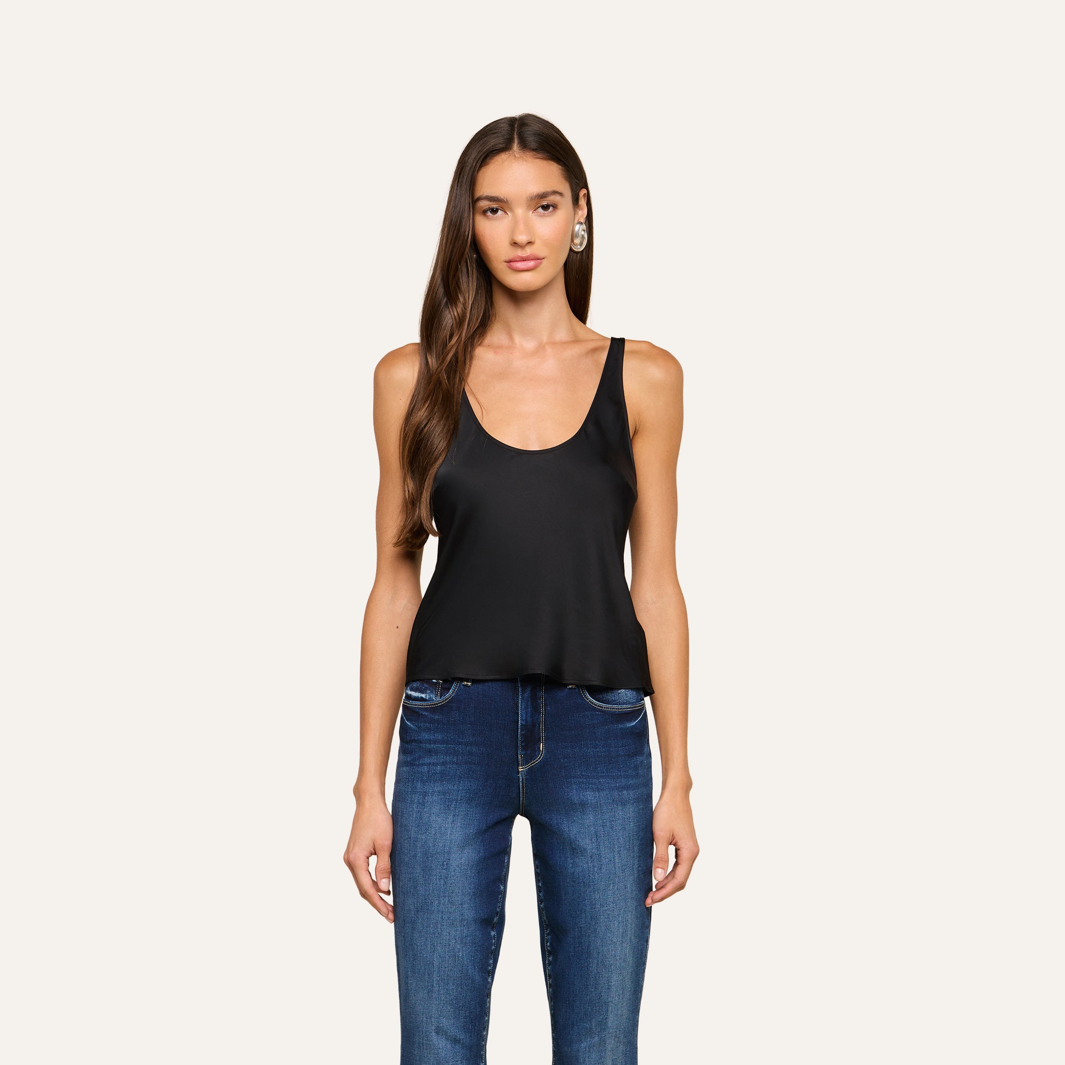 A woman with long brown hair stands facing forward against a plain light background, wearing the Eddie Tank—a black sleeveless top with a scoop neckline—paired with blue jeans.