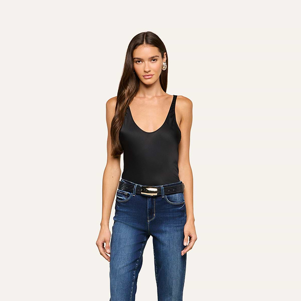 A woman with long brown hair is wearing the Eddie Tank—a sleeveless black bodysuit with a flattering drape—paired with blue jeans and a belt, standing against a plain light background.
