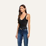A woman with long brown hair wears the Eddie Tank—a black scoop-neckline tank bodysuit—paired with blue jeans, a black belt featuring a gold buckle, and earrings, as she stands against a plain light background.