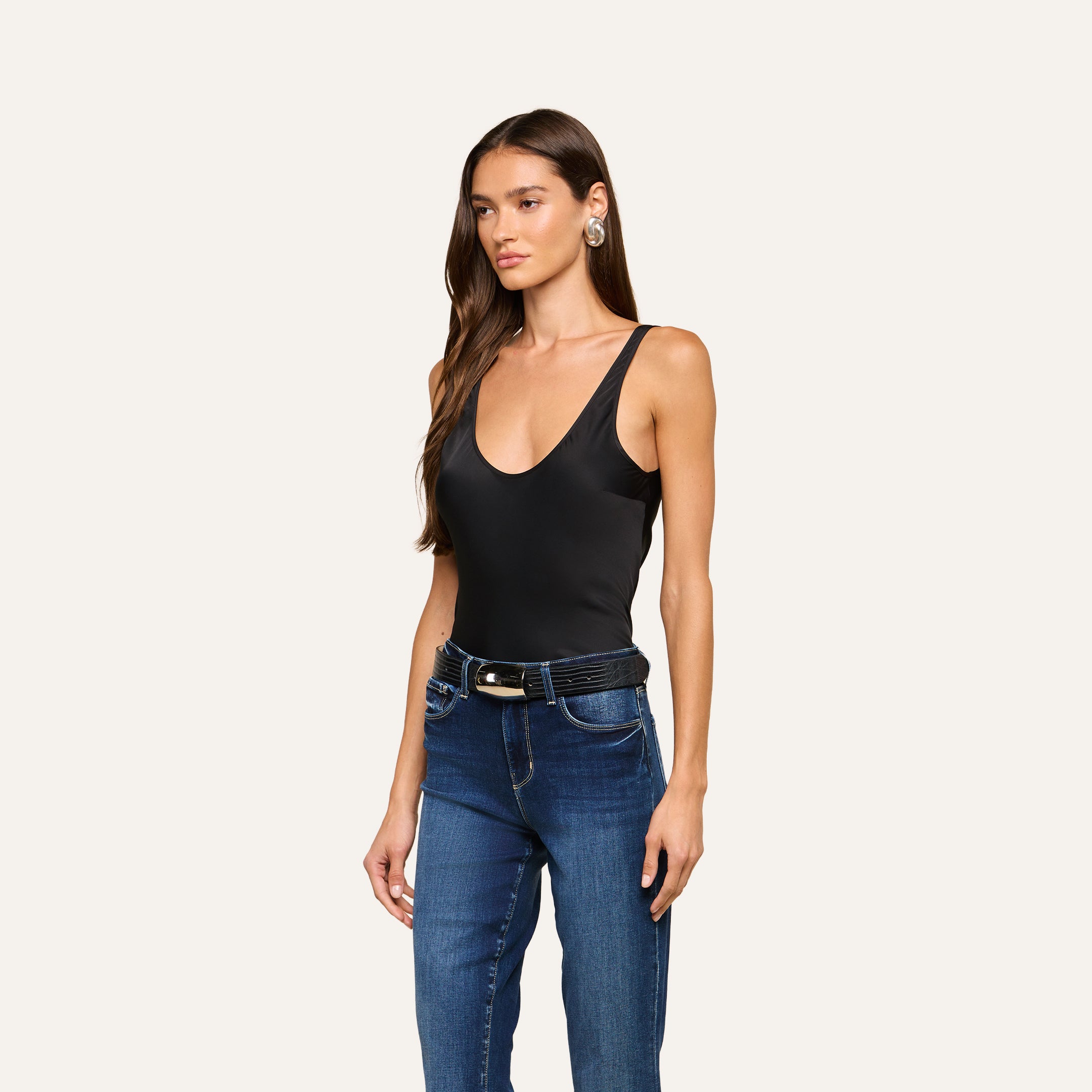 A woman with long brown hair wears the Eddie Tank—a black scoop-neckline tank bodysuit—paired with blue jeans, a black belt featuring a gold buckle, and earrings, as she stands against a plain light background.