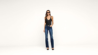 A woman wearing sunglasses, a black sleeveless top, blue jeans, and black heels stands against a plain white background with one hand in her pocket.