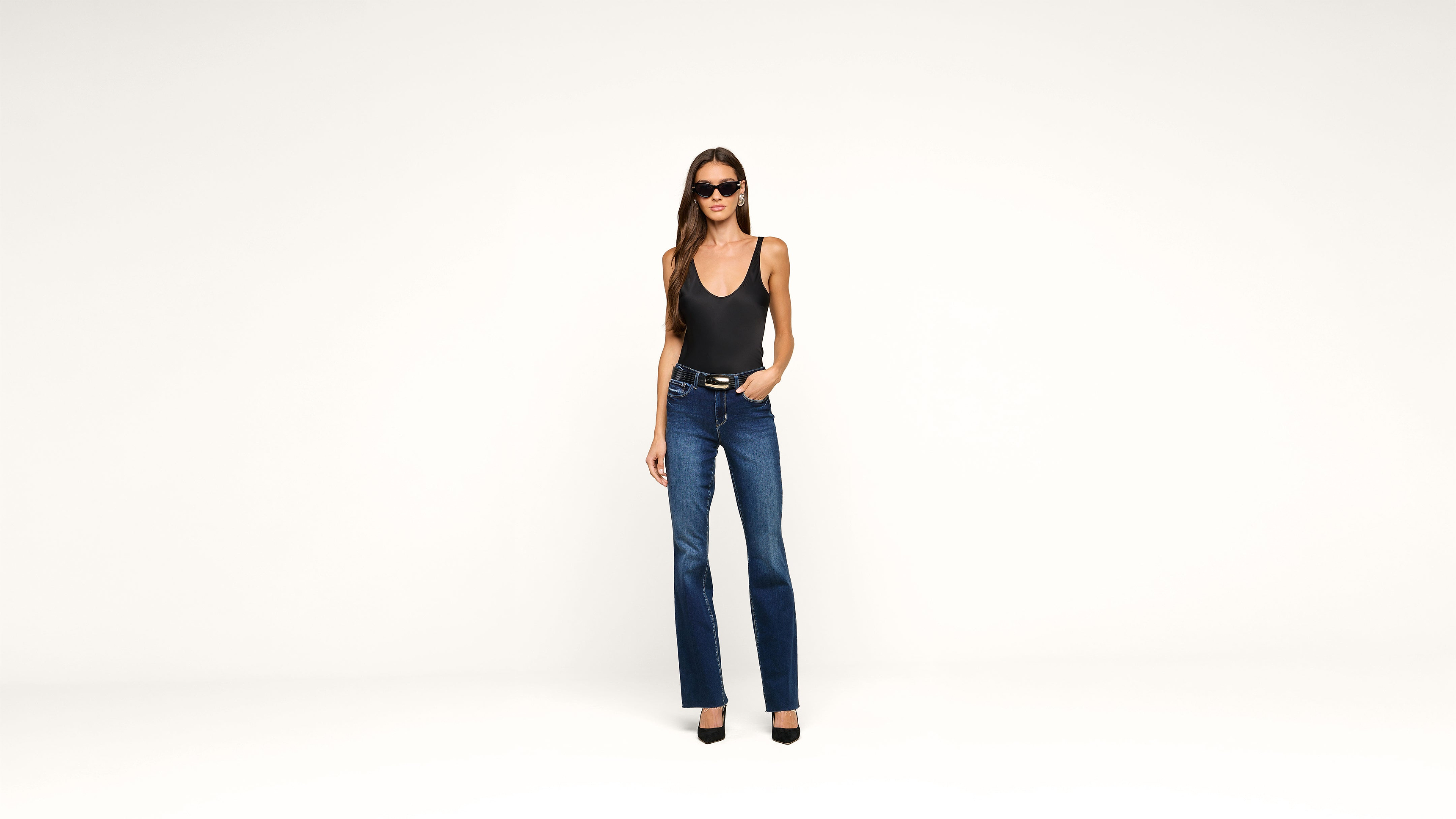 A woman wearing sunglasses, a black sleeveless top, blue jeans, and black heels stands against a plain white background with one hand in her pocket.