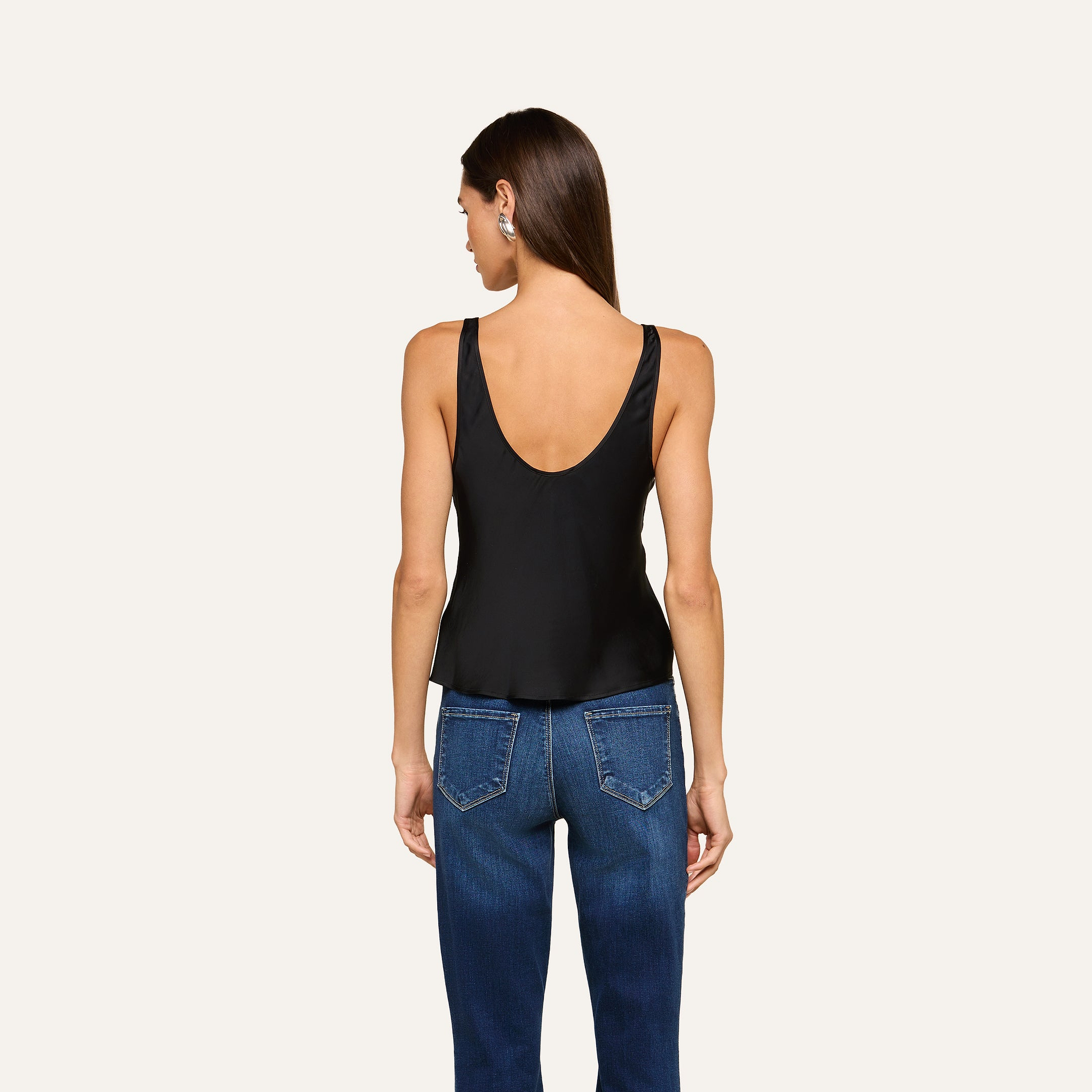 A woman with straight brown hair wears the Eddie Tank—a sleeveless black top with a flattering drape—paired with blue jeans, standing against a plain light background and facing away from the camera.