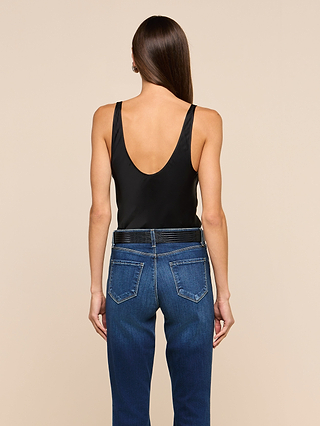 A woman with long brown hair stands against a beige background, seen from behind, wearing the Eddie Tank—a black bias-cut satin tank top with a deep scoop back—paired with blue jeans.