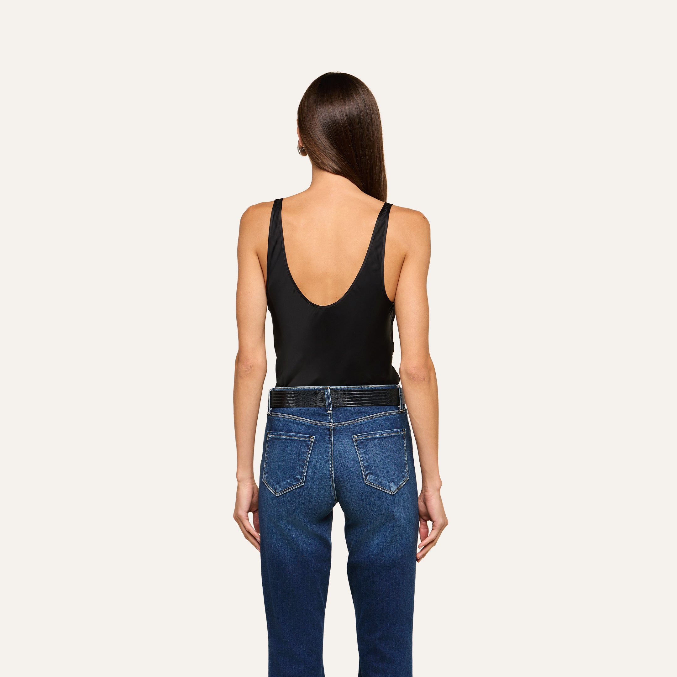 A person with long, straight brown hair is facing away, wearing the Eddie Tank—a black sleeveless top with a scoop neckline and flattering drape—paired with blue jeans, standing against a plain light background.
