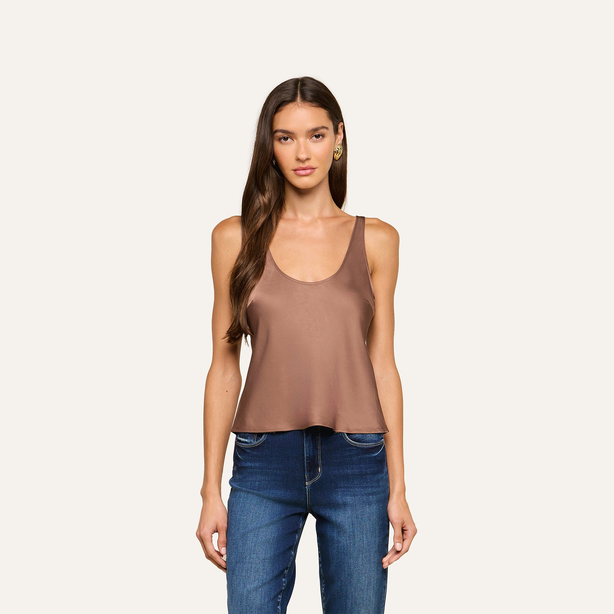 A woman with long brown hair wears the Eddie Tank, a sleeveless cardamom brown satin top with a scoop neckline, paired with blue jeans. She stands against a plain light background, facing forward with a neutral expression.