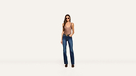 A woman wearing sunglasses, a brown tank top, blue jeans, and black boots stands against a plain white background with one hand in her pocket.