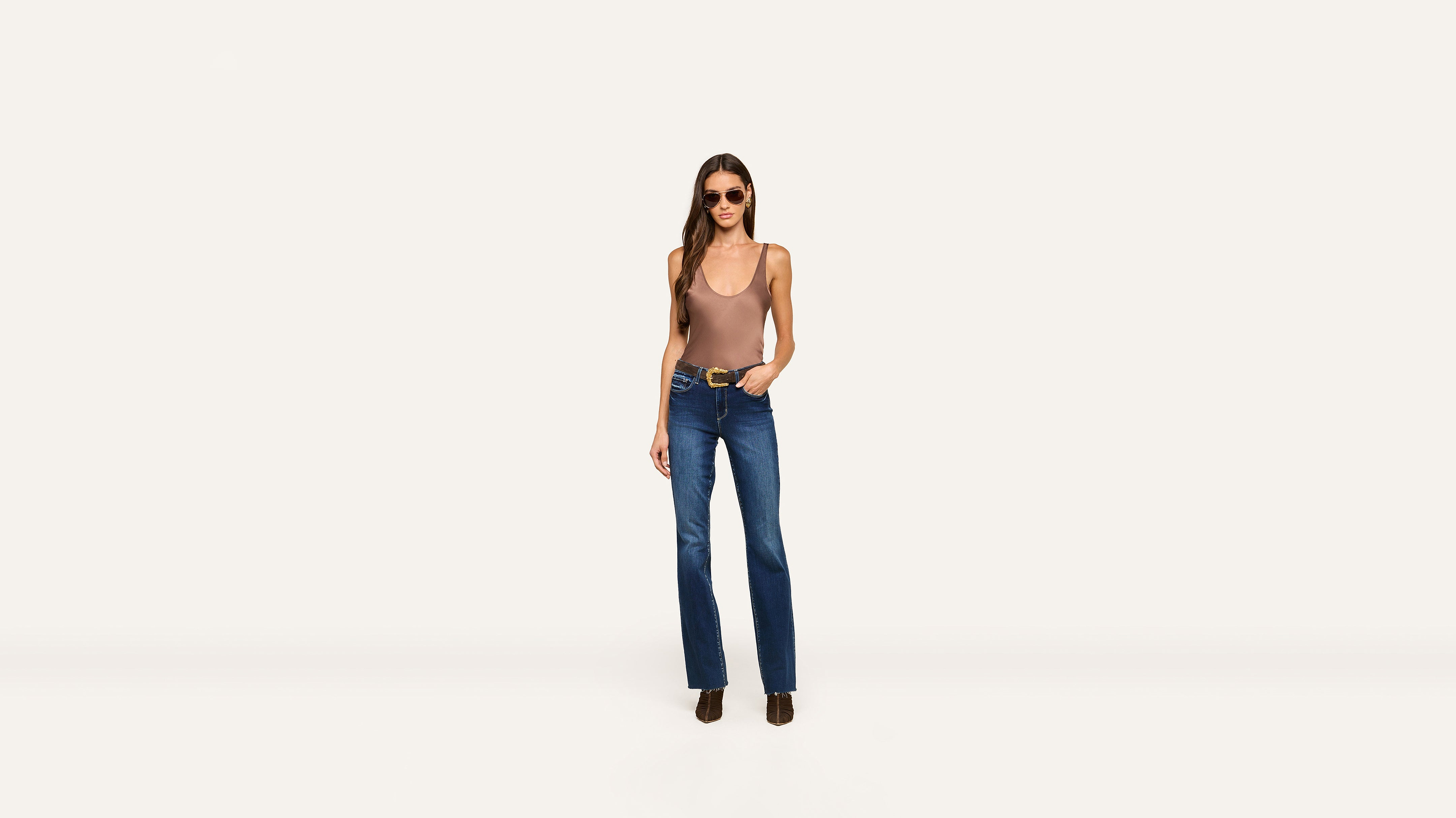 A woman wearing sunglasses, a brown tank top, blue jeans, and black boots stands against a plain white background with one hand in her pocket.