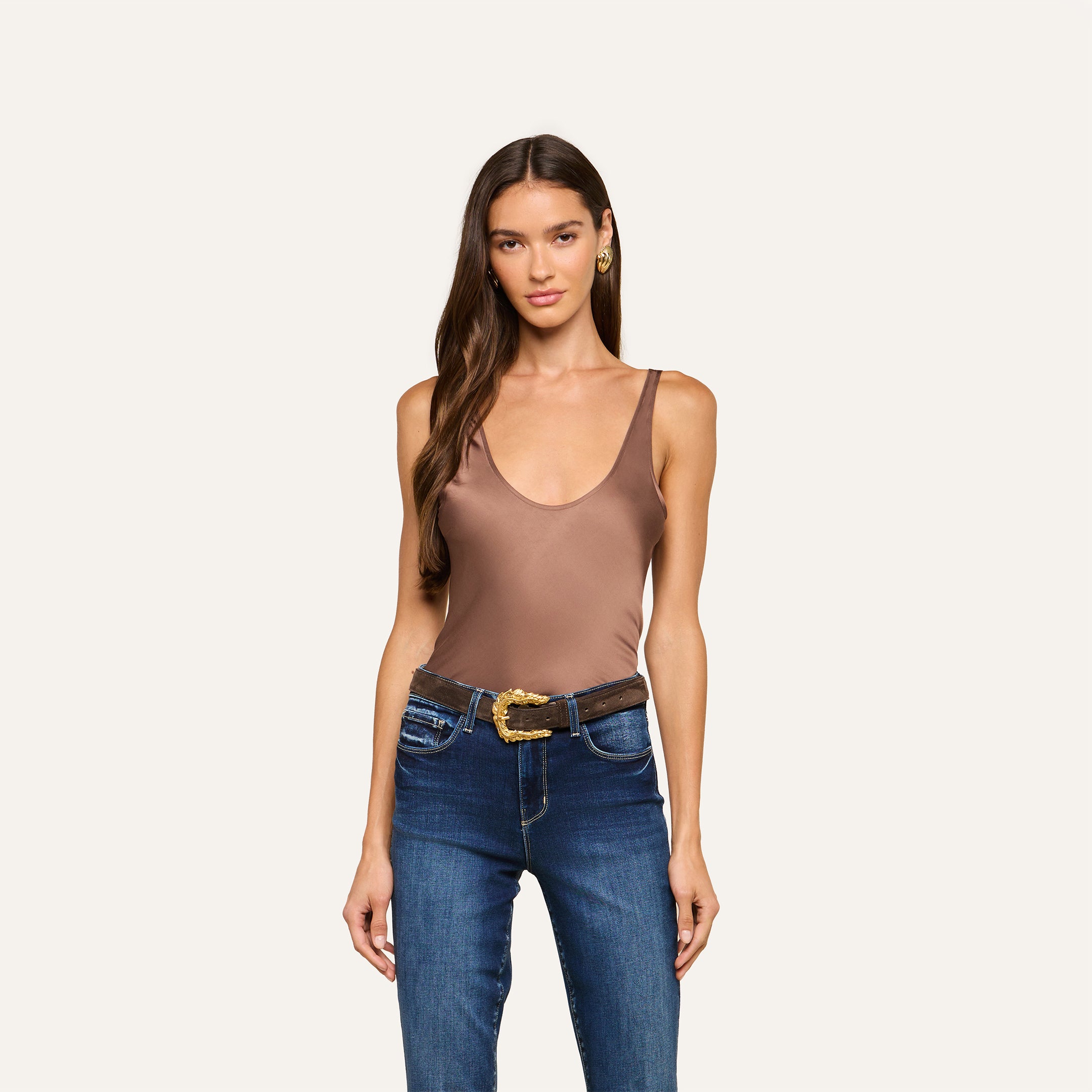 A woman with long brown hair wears the Eddie Tank, a bias-cut satin, cardamom brown sleeveless top with a scoop neckline, styled with blue jeans and a gold-buckled black belt, standing against a plain light background.