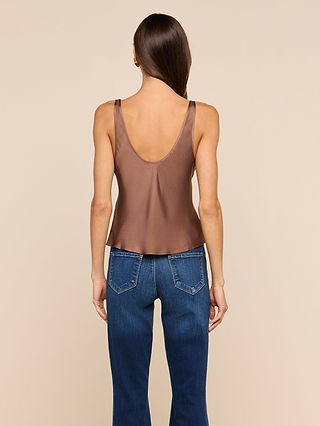 A woman stands facing away, wearing the Eddie Tank—a sleeveless cardamom brown bias-cut satin top with a low back—paired with blue jeans, against a beige background.