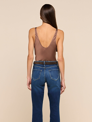 A woman stands facing away, wearing the Eddie Tank in cardamom brown—a sleeveless top featuring a low scoop neckline—paired with blue jeans. She has straight, long brown hair and is posed against a plain beige background.