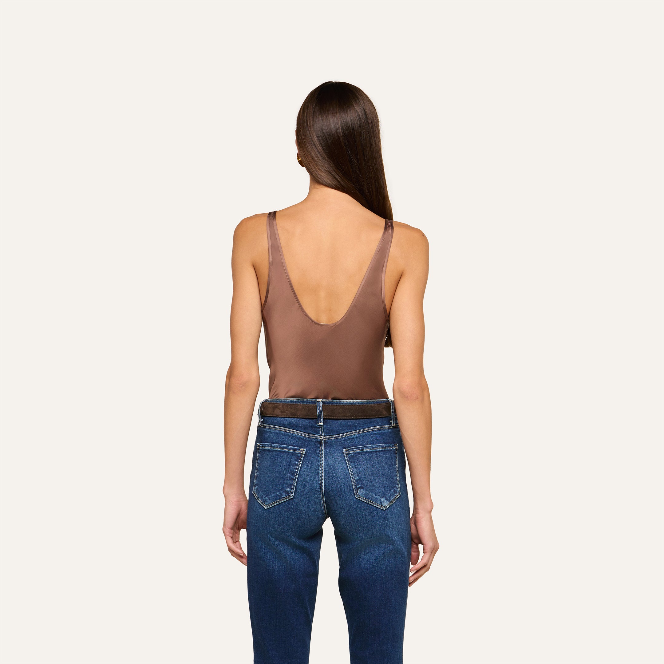 A woman with long straight brown hair stands with her back to the camera against a plain light background, wearing the Eddie Tank—a cardamom brown bias-cut satin sleeveless top with a low back—paired with blue jeans.