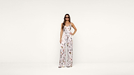 A woman wearing sunglasses and a white floral jumpsuit stands confidently with one hand on her hip against a plain white background.