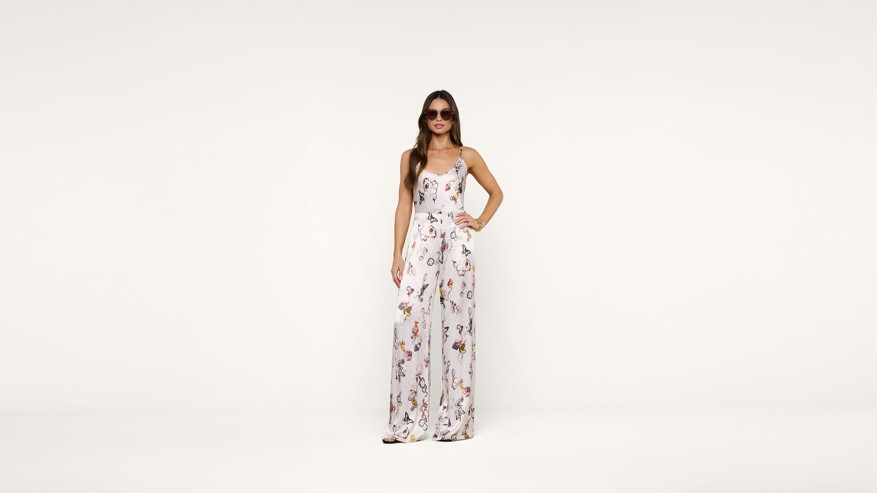 A woman wearing sunglasses and a white floral jumpsuit stands confidently with one hand on her hip against a plain white background.