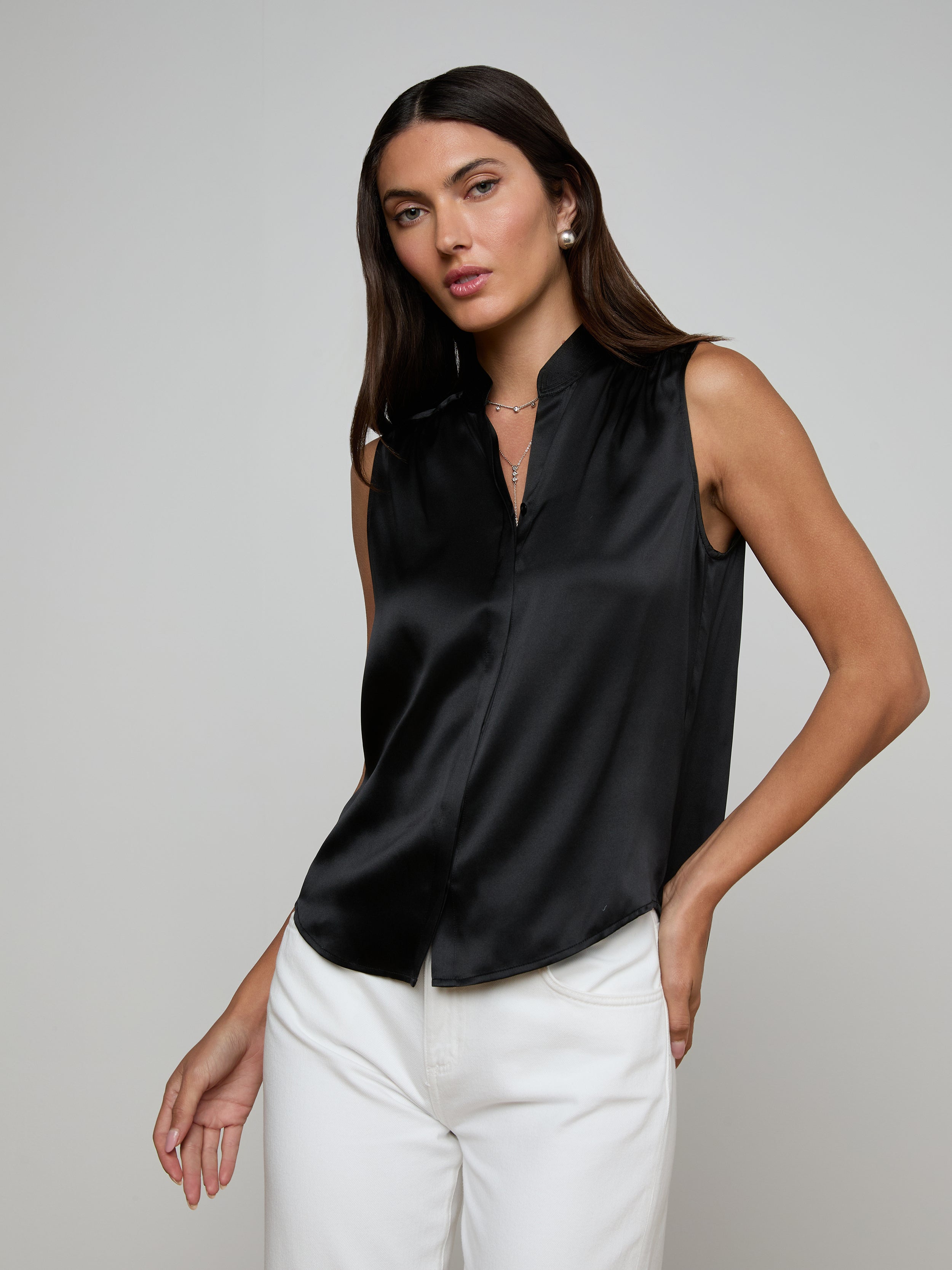A woman wearing the Phoebe Silk Blouse, a sleeveless black silk charmeuse top, paired with white pants stands against a plain light background, looking at the camera with one hand resting in her pocket.