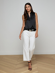 Wearing the Phoebe Silk Blouse—a sleeveless black silk charmeuse top—paired with white pants and black heels, a woman stands against a plain, light background, looking forward with a neutral expression.