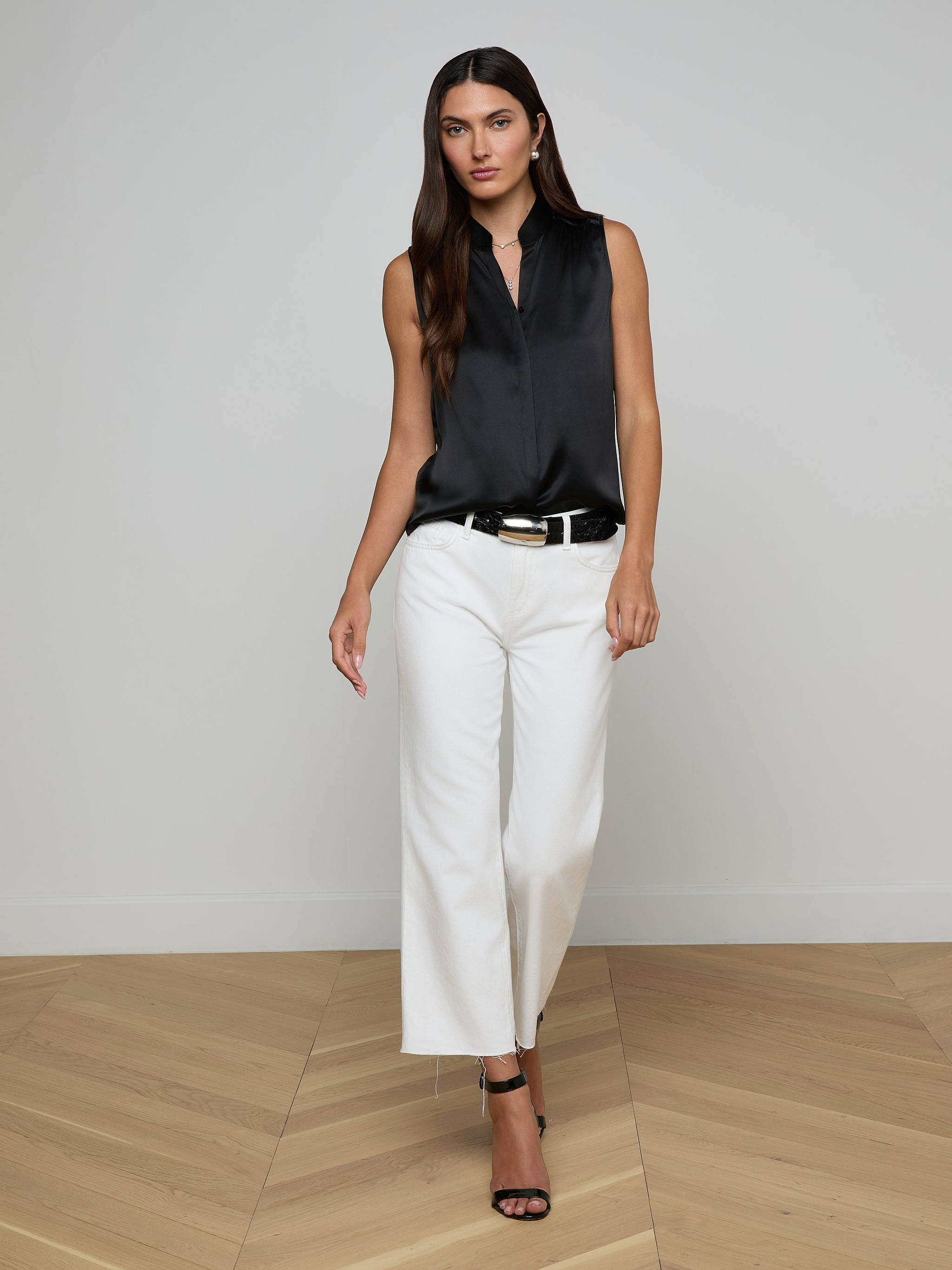 Wearing the Phoebe Silk Blouse—a sleeveless black silk charmeuse top—paired with white pants and black heels, a woman stands against a plain, light background, looking forward with a neutral expression.