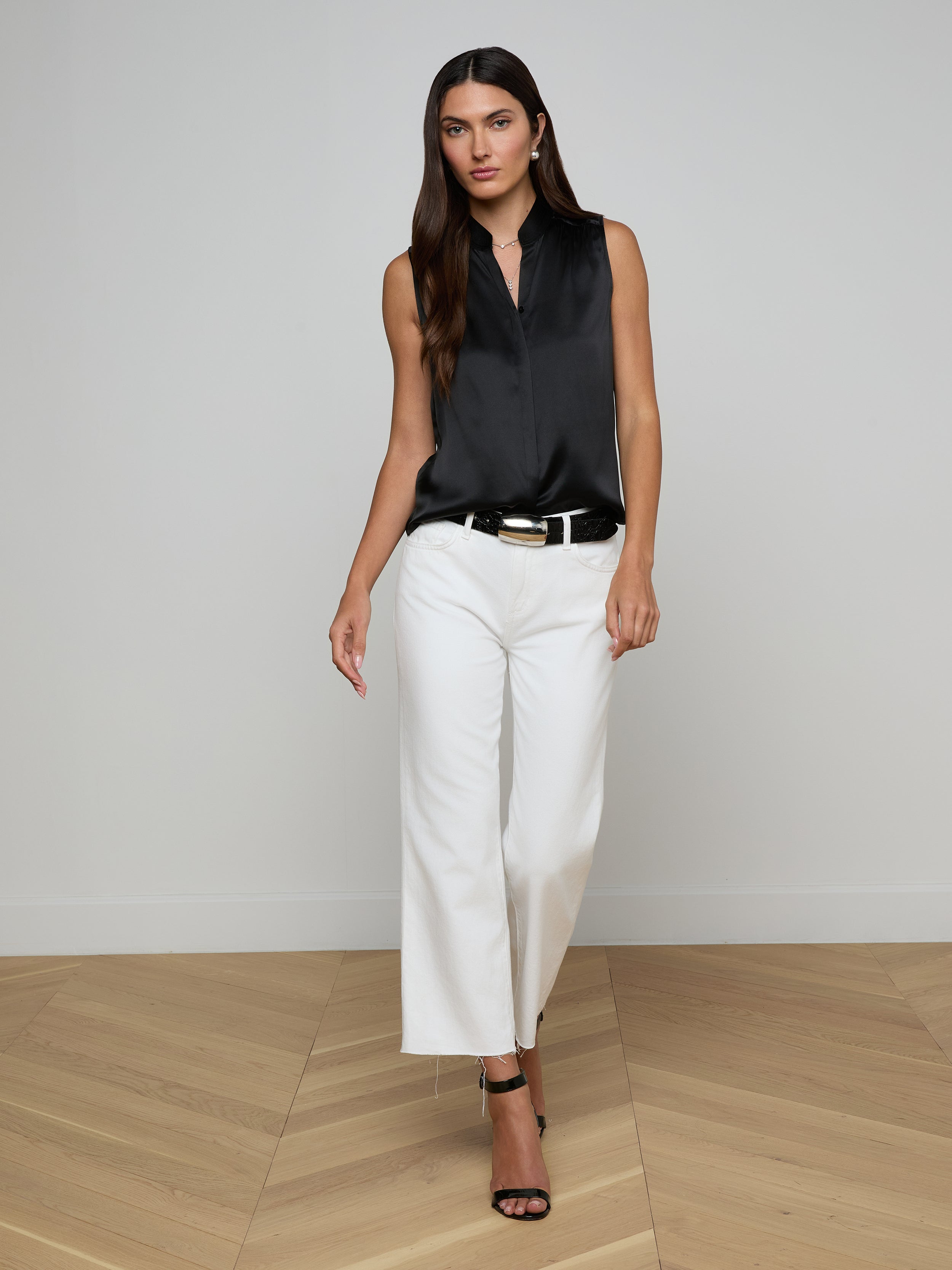 Wearing the Phoebe Silk Blouse—a sleeveless black silk charmeuse top—paired with white pants and black heels, a woman stands against a plain, light background, looking forward with a neutral expression.