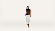A woman with long dark hair wears a sleeveless black blouse, white pants, and black heels. She stands against a plain white background, facing forward with a confident expression.