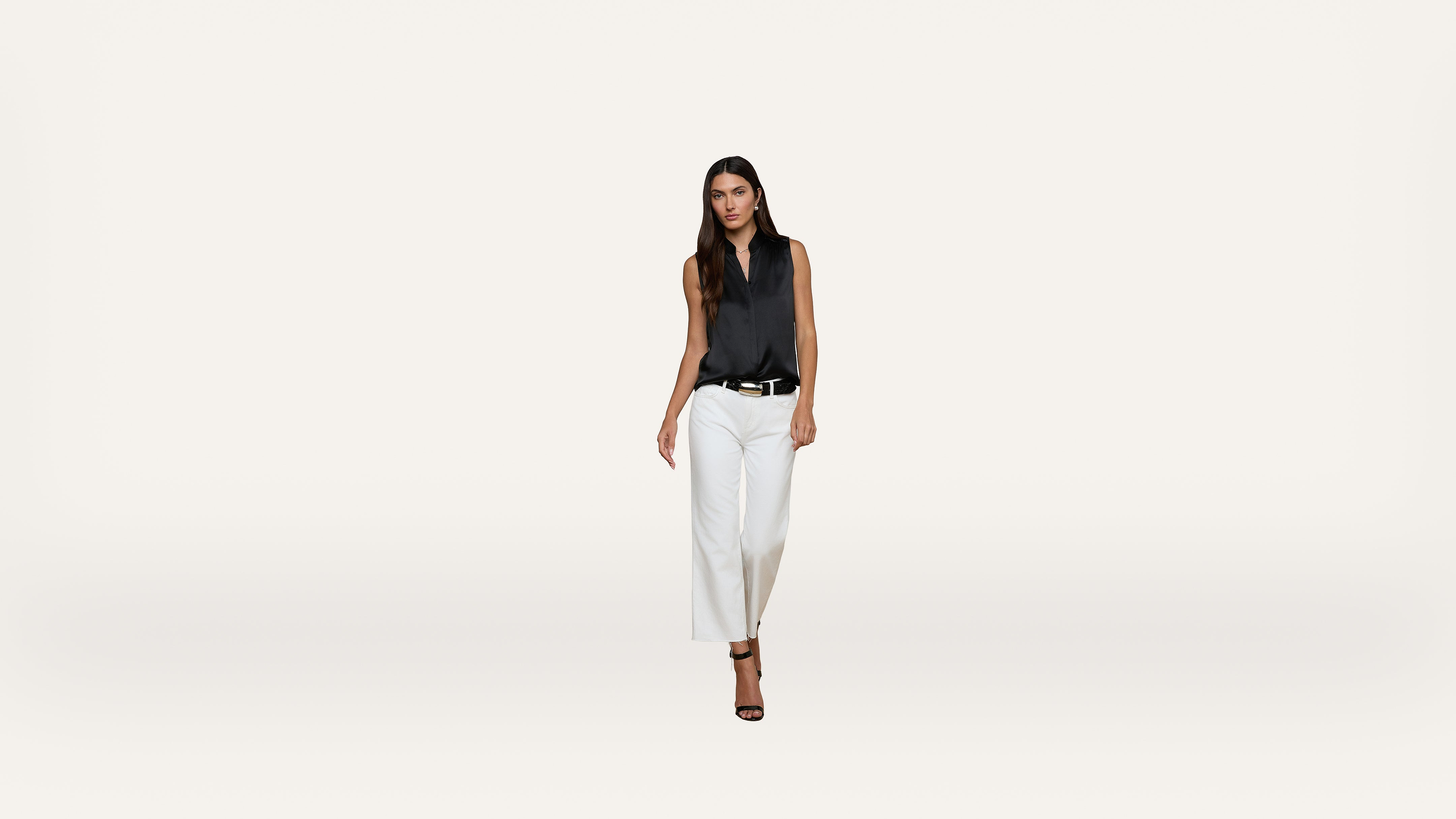 A woman with long dark hair wears a sleeveless black blouse, white pants, and black heels. She stands against a plain white background, facing forward with a confident expression.