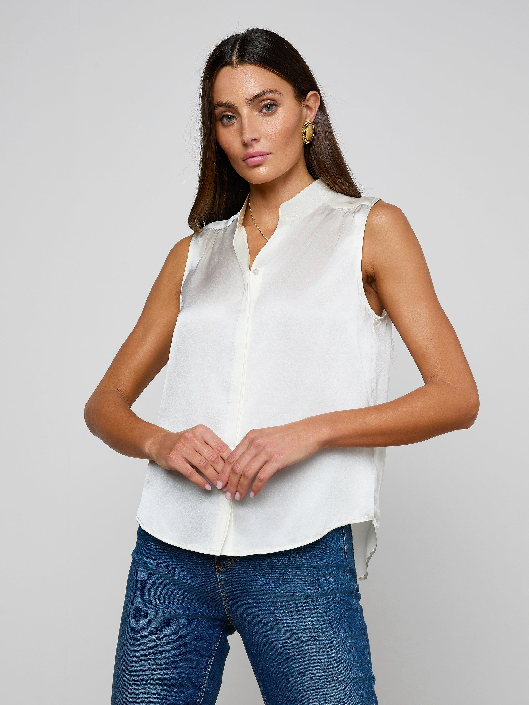 A woman with long brown hair wears the Phoebe Silk Blouse, a sleeveless silk charmeuse top, paired with blue jeans. She stands against a plain light background, looking at the camera with her hands gently clasped.