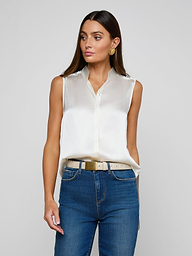 A woman wearing the Phoebe Silk Blouse in white, paired with blue jeans and a beige belt, stands against a plain light background, looking slightly to the side. She has long brown hair and accessorizes with gold hoop earrings.