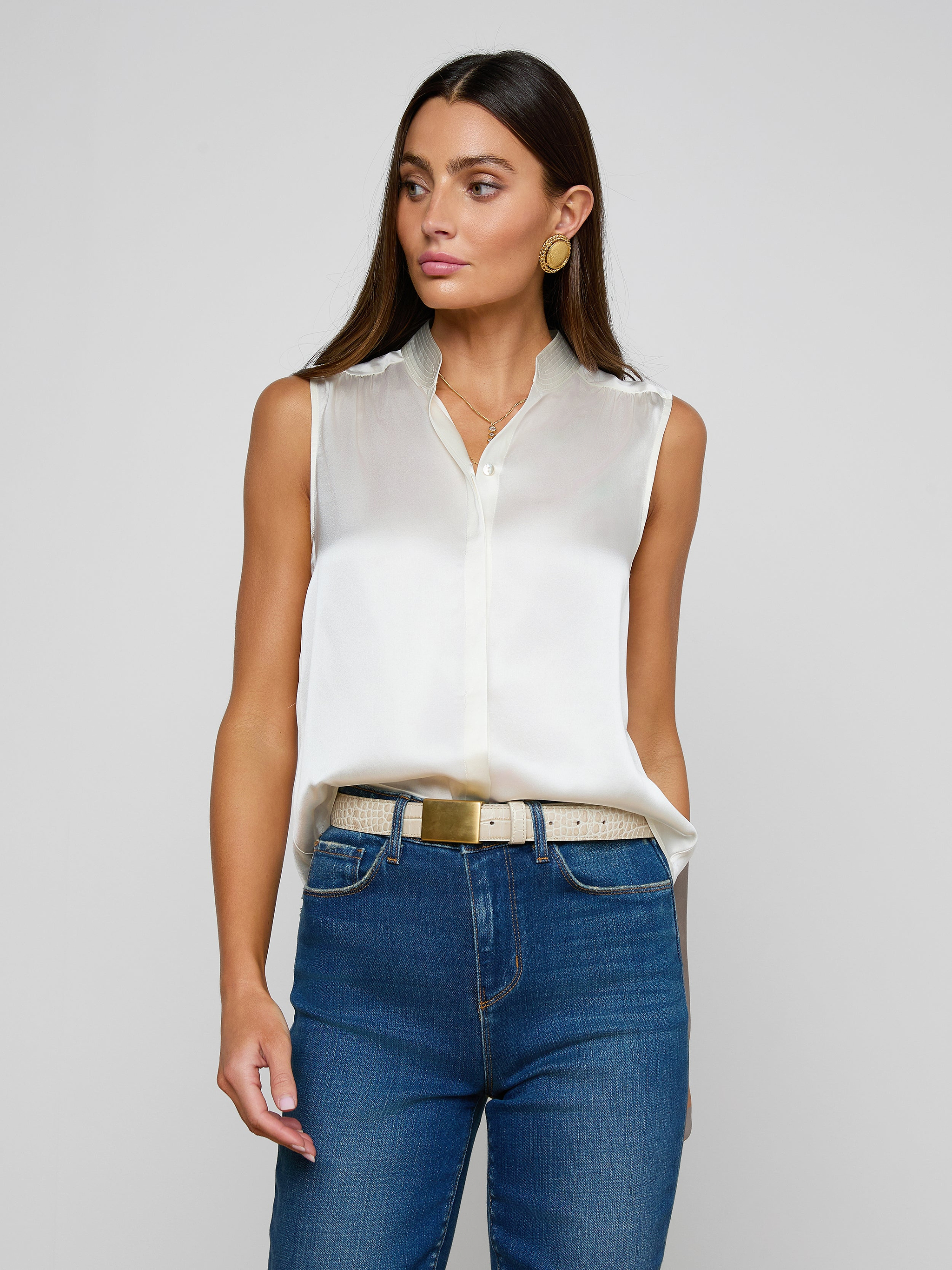 A woman wearing the Phoebe Silk Blouse in white, paired with blue jeans and a beige belt, stands against a plain light background, looking slightly to the side. She has long brown hair and accessorizes with gold hoop earrings.