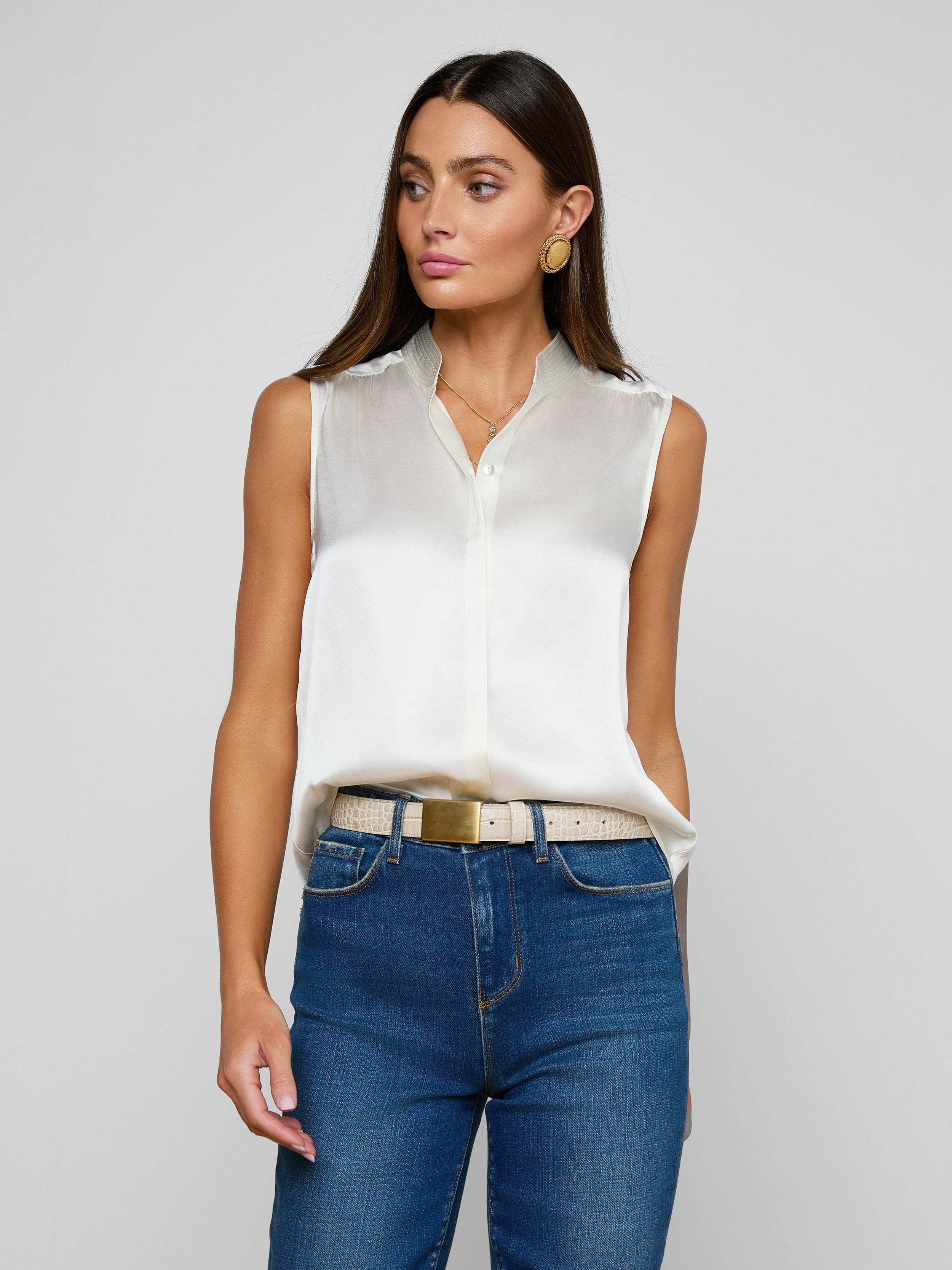 A woman wearing the Phoebe Silk Blouse in white, paired with blue jeans and a beige belt, stands against a plain light background, looking slightly to the side. She has long brown hair and accessorizes with gold hoop earrings.