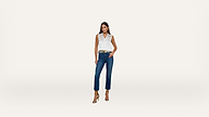 A woman stands against a plain white background, wearing a sleeveless white top, blue jeans, a brown belt, and beige high-heeled sandals. One hand is in her pocket and the other rests by her side.