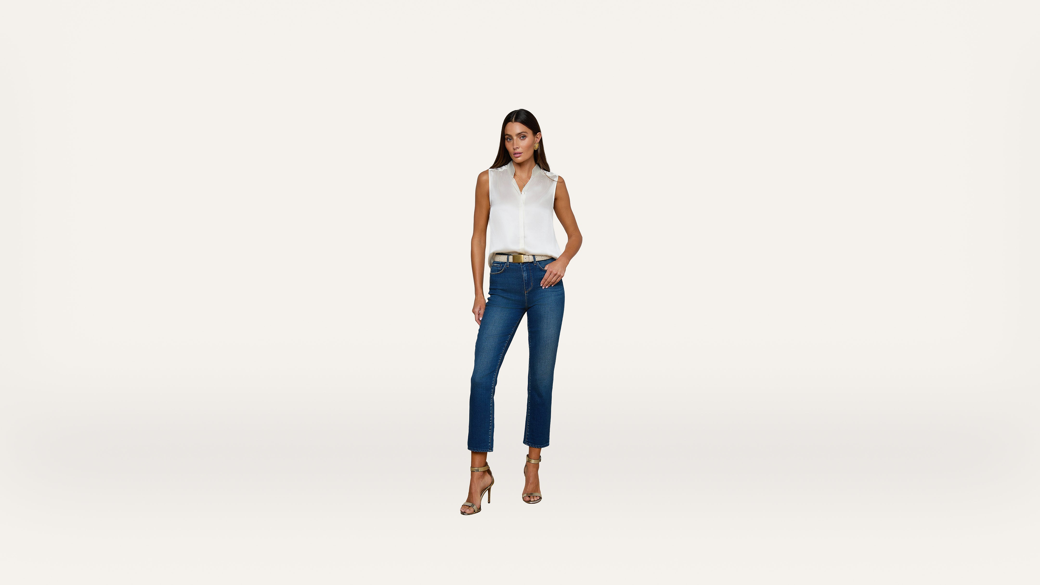 A woman stands against a plain white background, wearing a sleeveless white top, blue jeans, a brown belt, and beige high-heeled sandals. One hand is in her pocket and the other rests by her side.