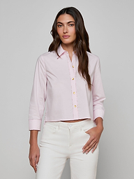 A woman models the Fawn Shirt, a cropped pale pink top, paired with white pants as she stands against a plain, light background with one hand in her pocket.
