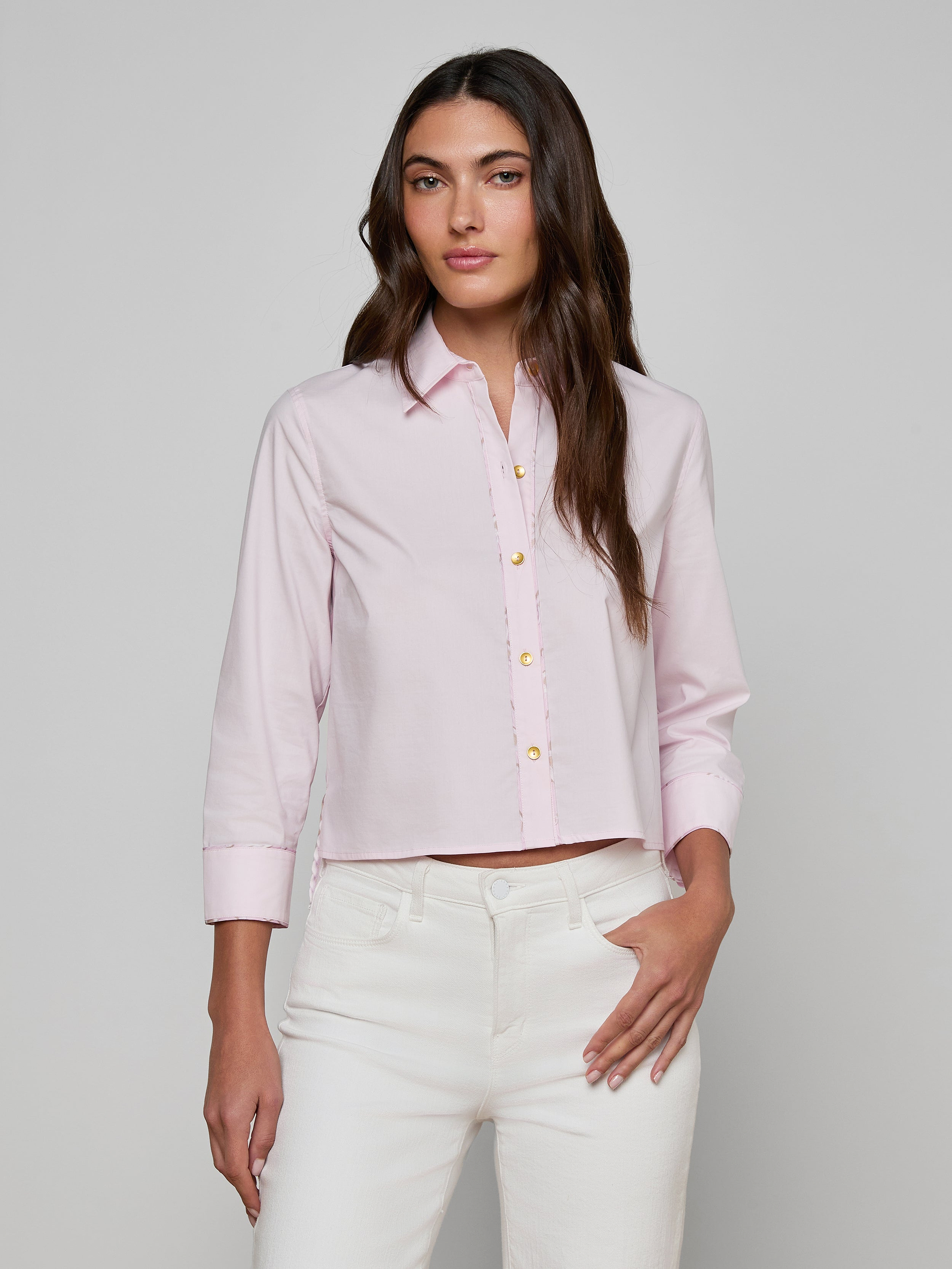 A woman models the Fawn Shirt, a cropped pale pink top, paired with white pants as she stands against a plain, light background with one hand in her pocket.
