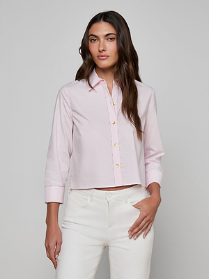 A woman with long brown hair wears the Fawn Shirt—an organic cotton blouse in light pink—and white pants, standing against a plain light gray background with one hand in her pocket and a neutral expression.