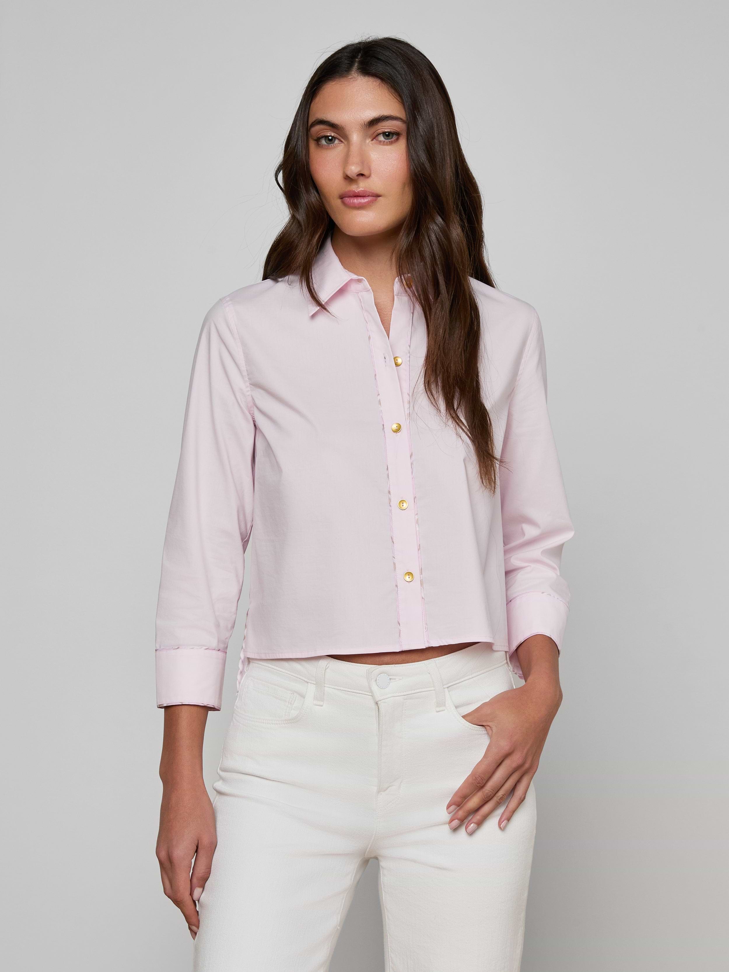 A woman models the Fawn Shirt, a cropped pale pink top, paired with white pants as she stands against a plain, light background with one hand in her pocket.