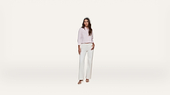 A woman stands against a plain white background, wearing a light pink long-sleeve button-up shirt, white wide-leg pants, and sandals.