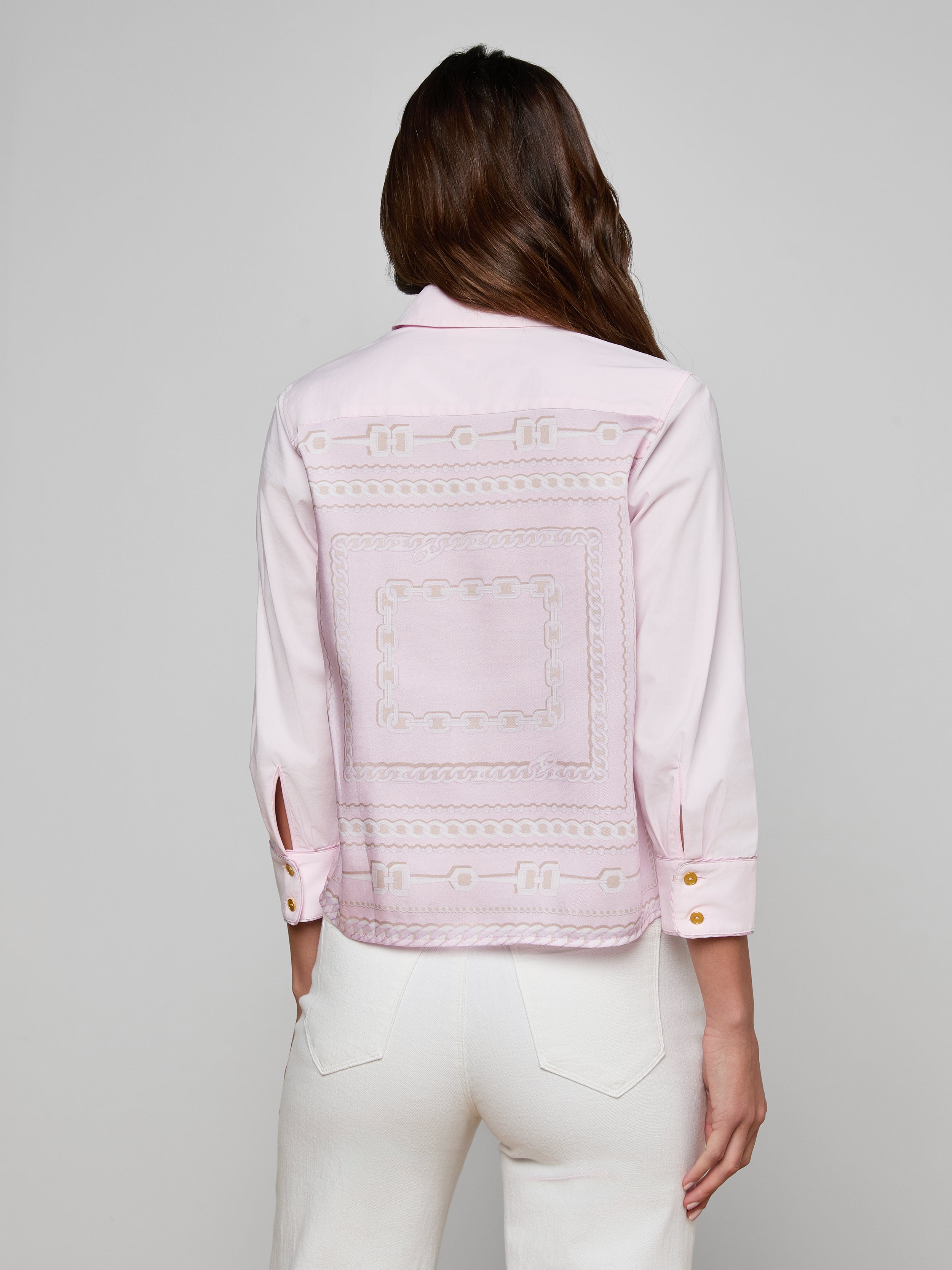 A woman with long brown hair wears the Fawn Shirt, a cropped pale pink top with an embroidered square design on the back, paired with white pants. She stands facing away against a plain, off-white background.