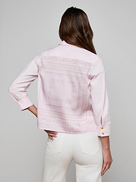 A woman with long brown hair, standing with one hand in her pocket and facing away from the camera against a plain background, wears the Fawn Shirt—a cropped pale pink top featuring embroidered patterns on the back—paired with white pants.