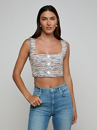 A woman wearing the Muno Tweed Top—a sleeveless, cropped style with frayed edges and large buttons—paired with light blue high-waisted jeans, stands against a plain light background.