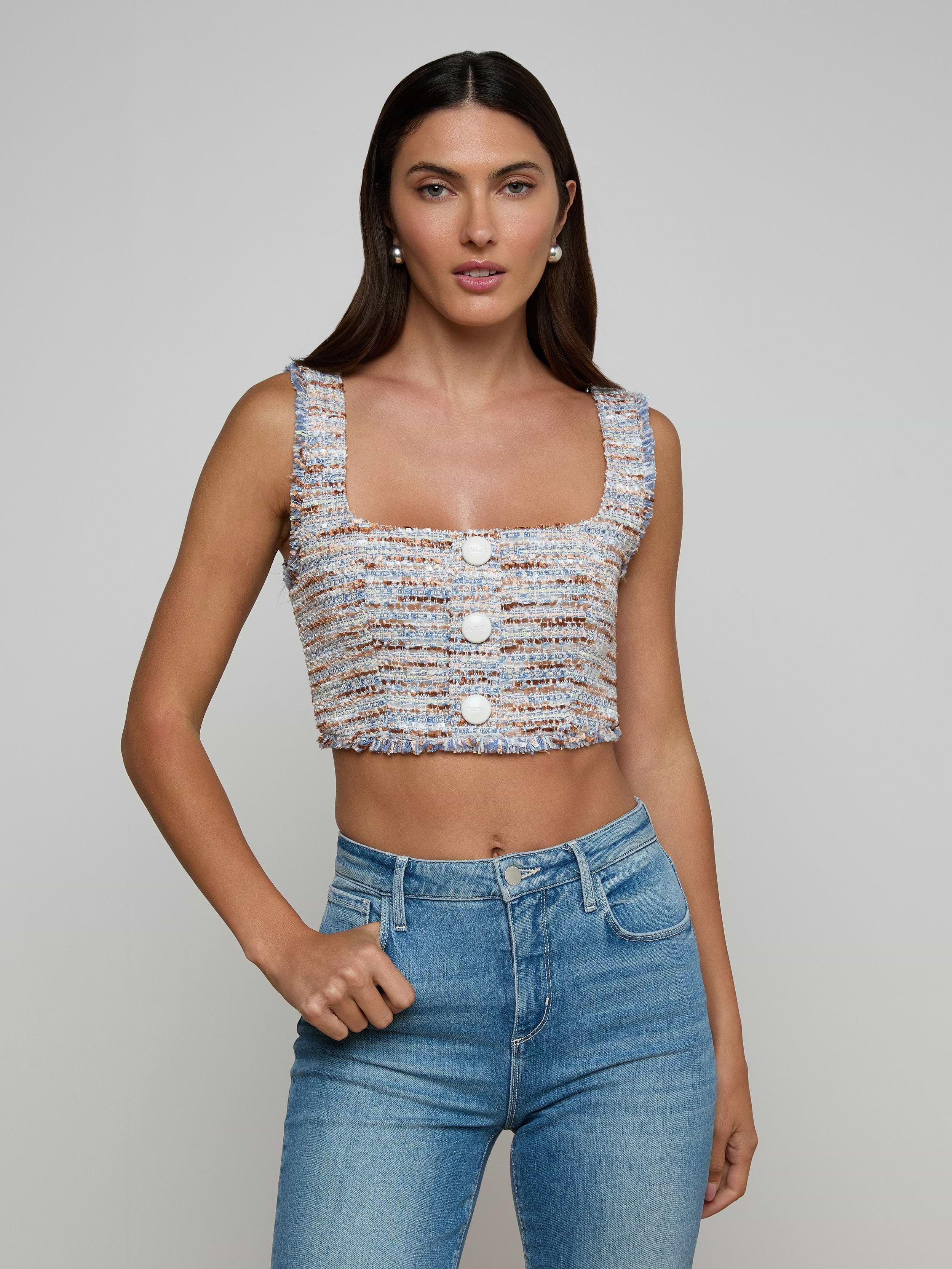 A woman wearing the Muno Tweed Top—a sleeveless, cropped style with frayed edges and large buttons—paired with light blue high-waisted jeans, stands against a plain light background.