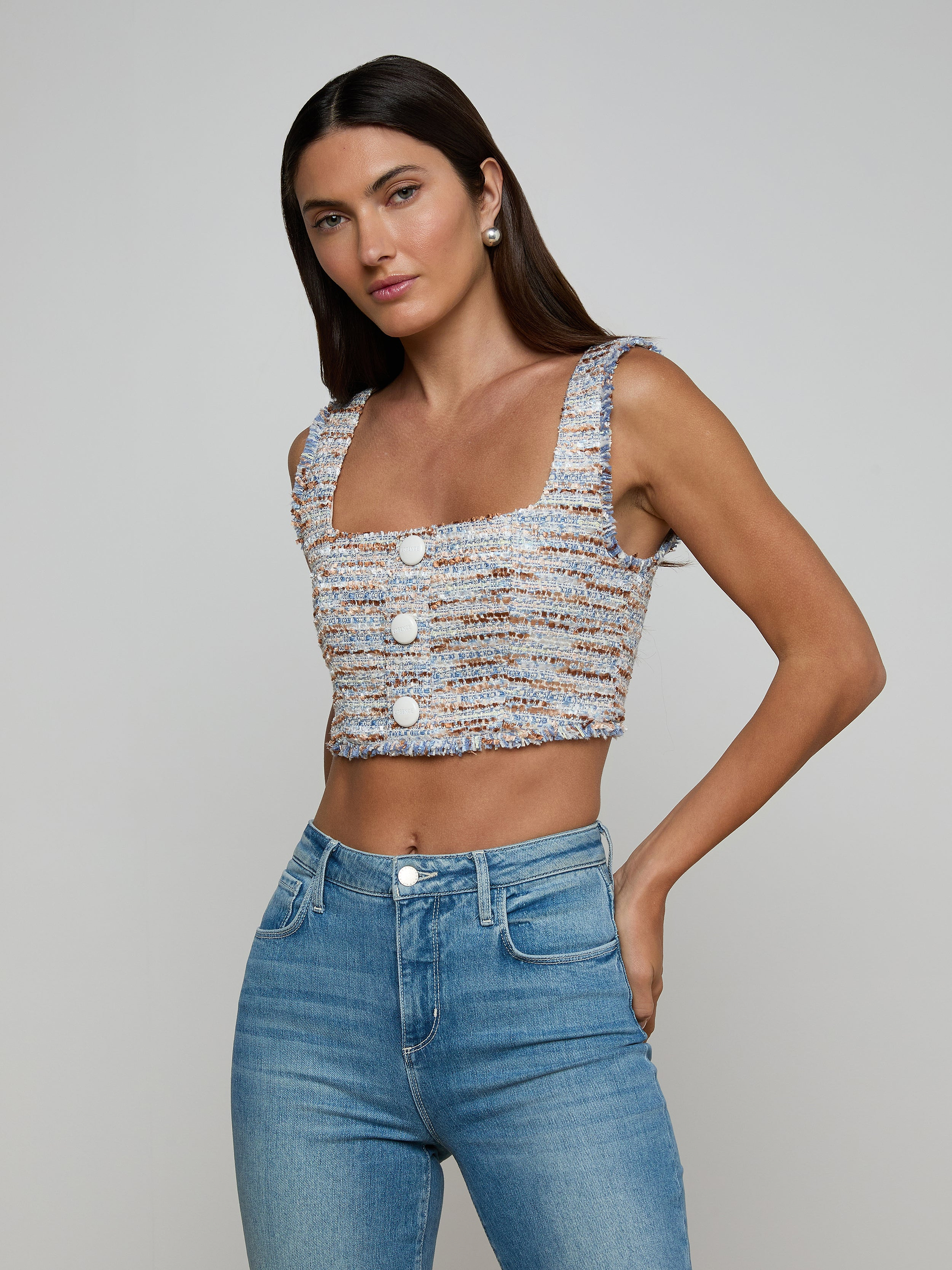 A woman stands with one hand on her hip against a plain beige background, wearing the Muno Tweed Top—a sleeveless, textured cropped top with large buttons—paired with high-waisted blue jeans.