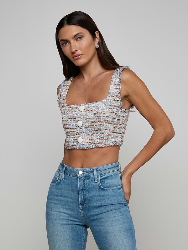 A woman with long dark hair wears the Muno Tweed Top—a sleeveless, square-neck, button-front crop with frayed edges—paired with high-waisted blue jeans, standing against a plain light gray background.