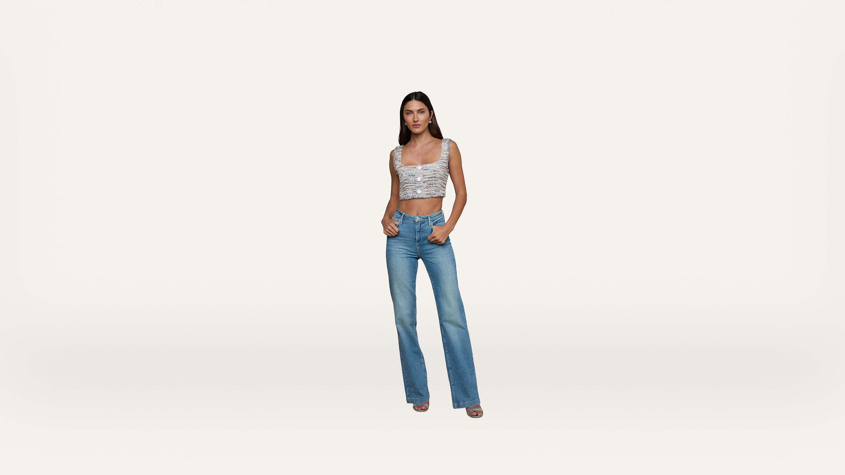 A woman stands against a plain white background, wearing a patterned crop top, high-waisted blue jeans, and heeled sandals, with one hand in her pocket and a neutral expression.