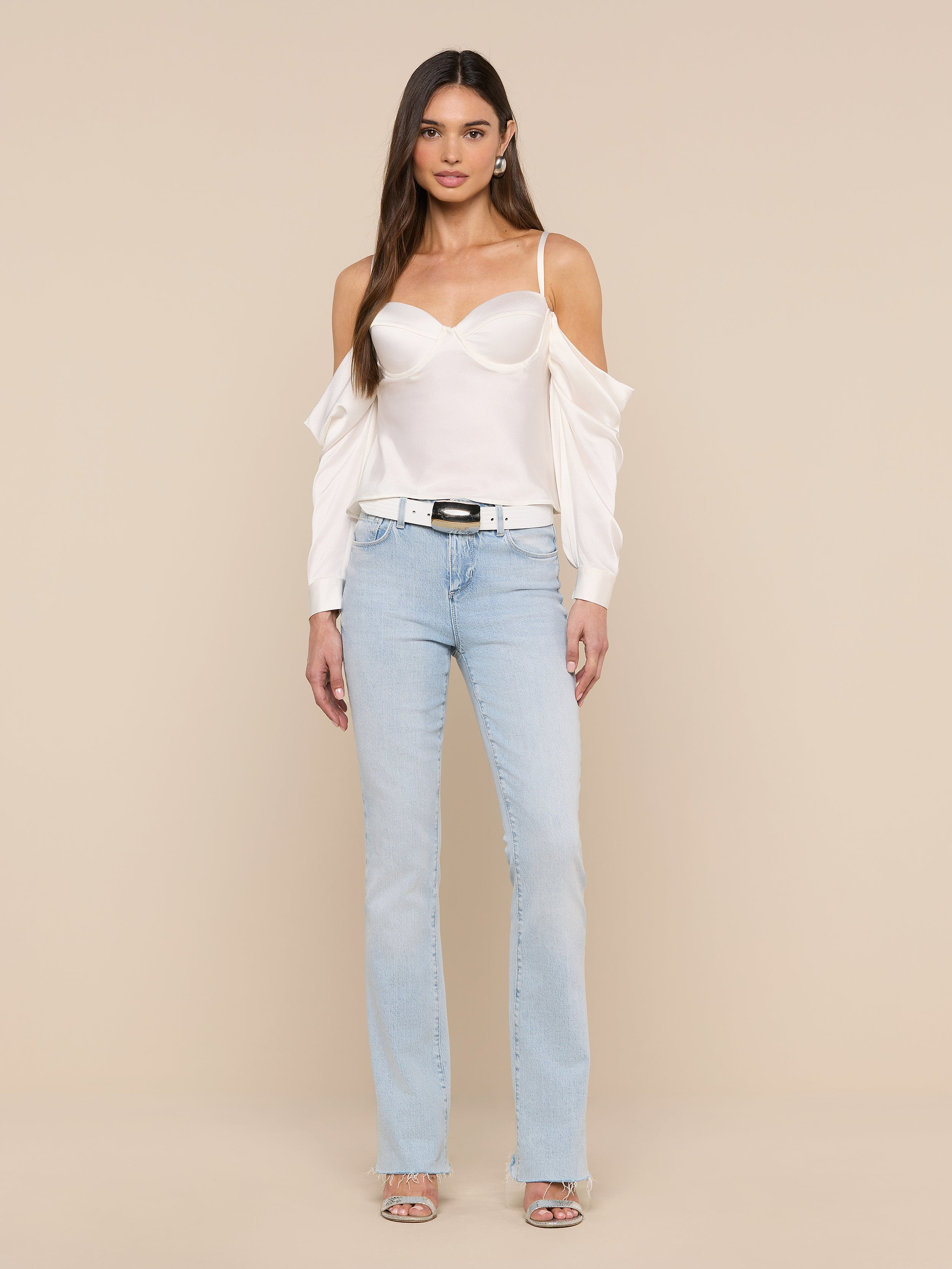 A woman stands against a plain white background wearing the Saya Satin Top in white, light blue jeans, a black belt, and open-toe heels. Her arms are relaxed by her sides and her long, straight hair is loose.